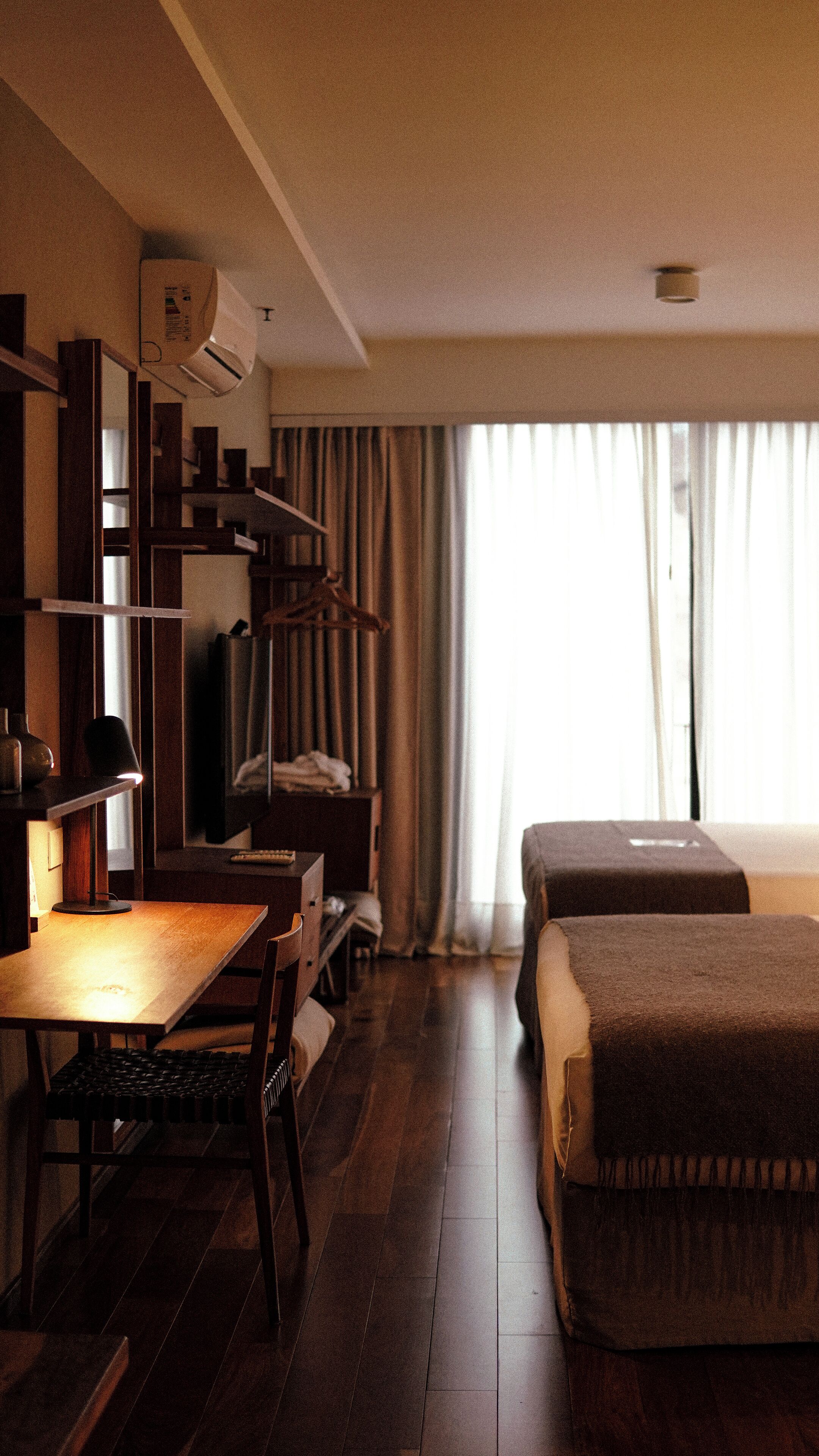 A hotel room with a wooden desk, a chair, a bed, a TV, a bookshelf, and a window with curtains.