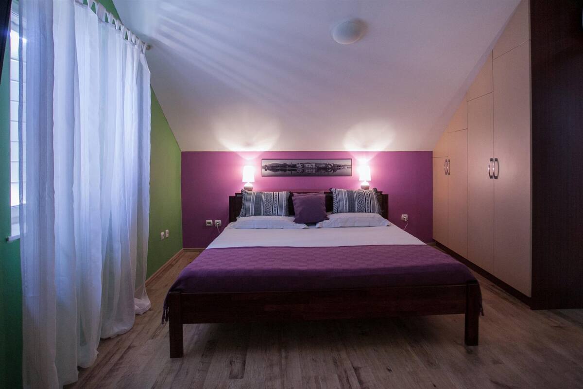 A bedroom with a large bed, two bedside tables with lamps, a wall-mounted air conditioner, and a green accent wall.