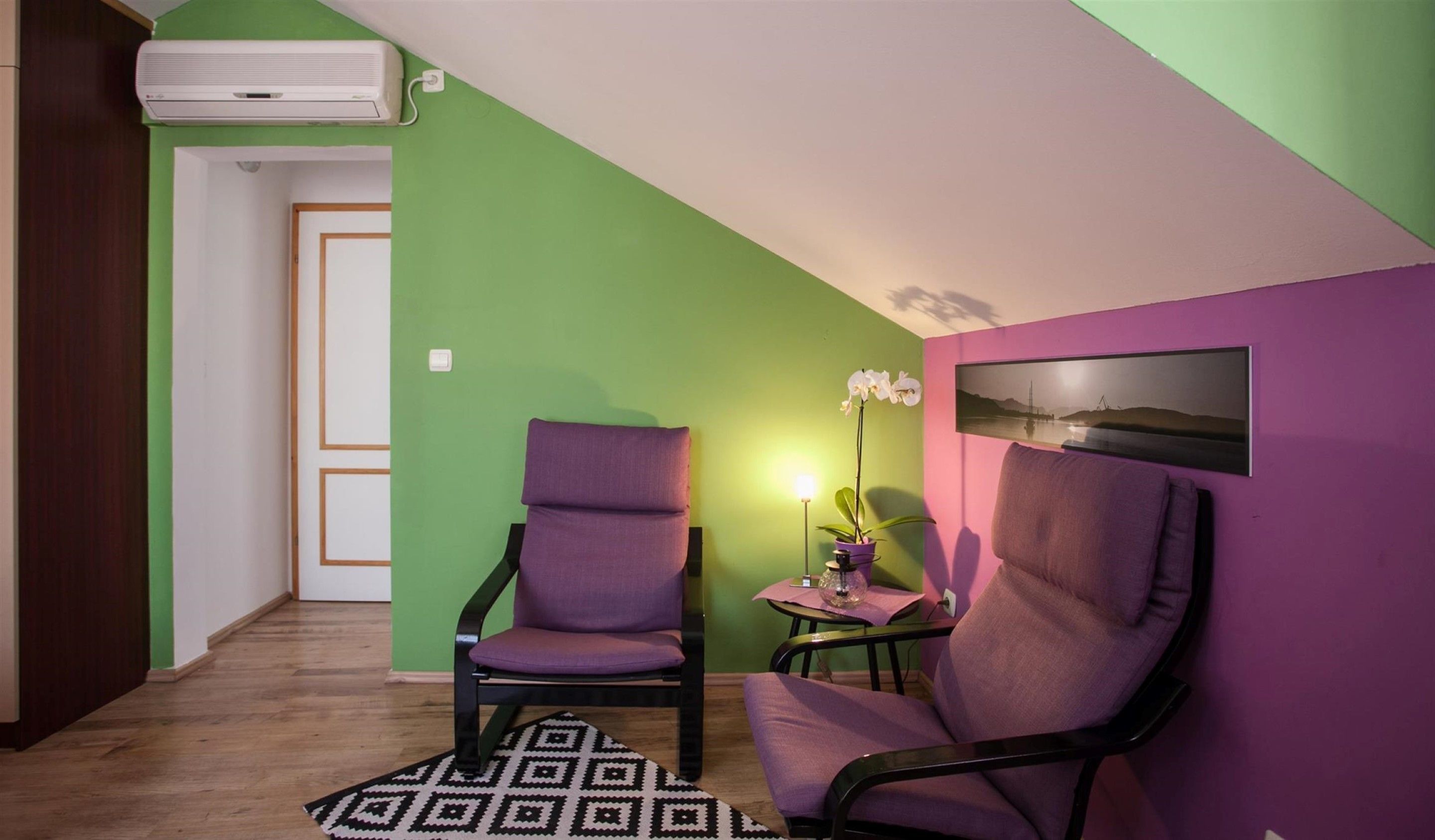 A room with two purple chairs, a small table with a potted orchid, a framed picture on the wall, and an air conditioning unit on the ceiling.