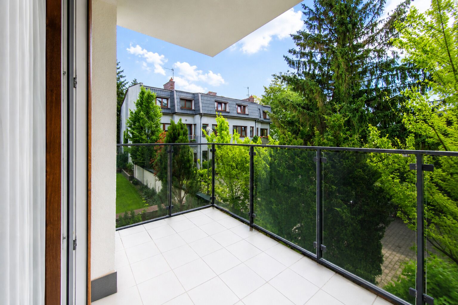 A balcony with a glass railing overlooking a residential area with trees and a house.
