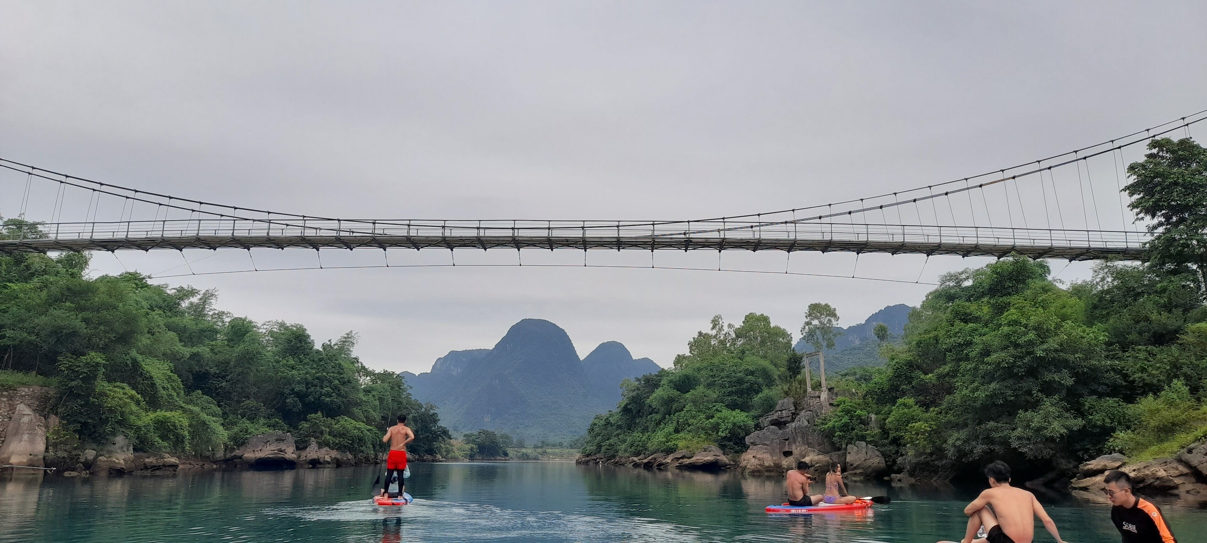 A river with people paddleboarding, a bridge overhead, and lush greenery in the background.