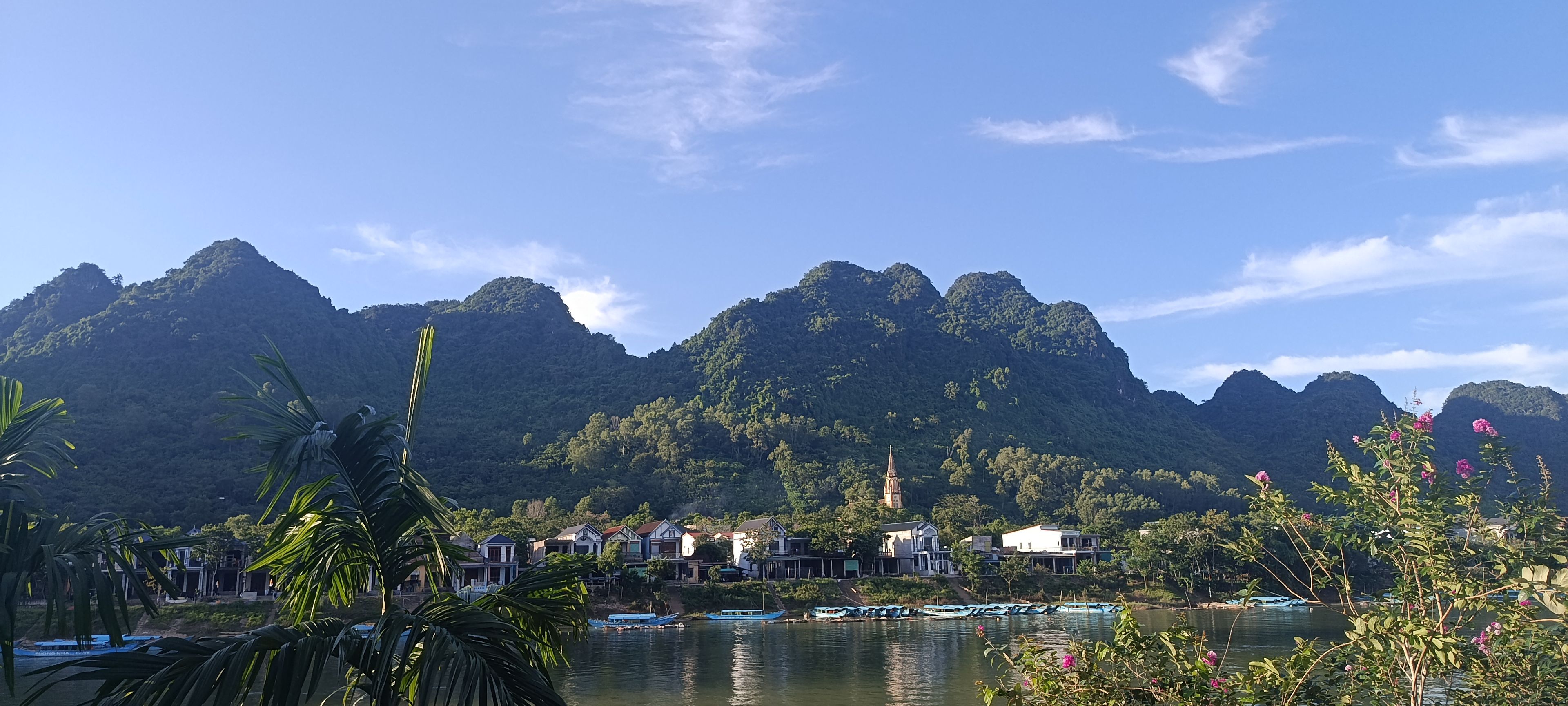 Vista sobre un lago con montañas al fondo, barcos amarrados a la orilla y un conjunto de edificios con una torre destacada.