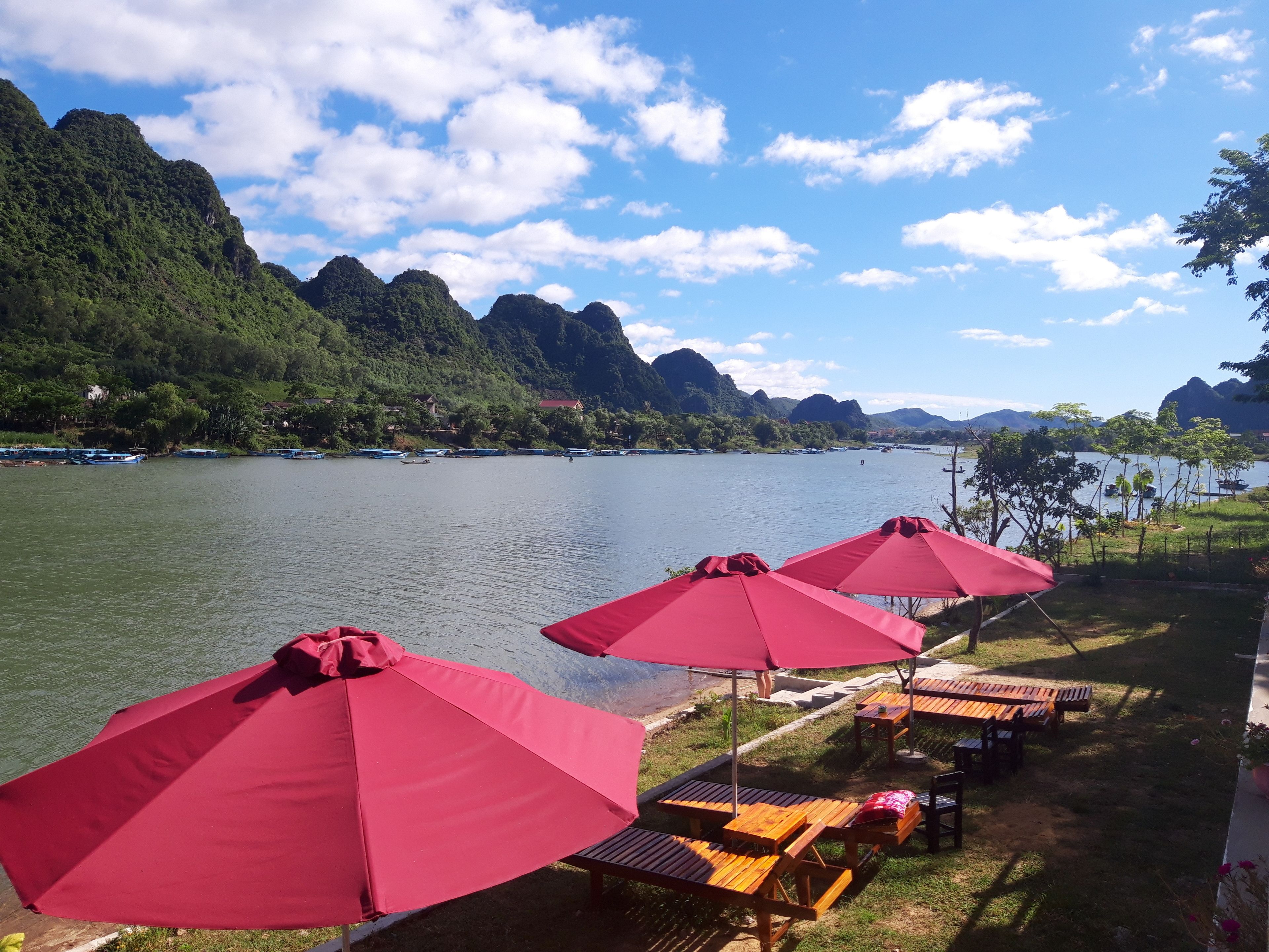 Une vue sur le lac, avec des parasols rouges et des tables en bois, le tout entouré d’une végétation luxuriante et de montagnes.