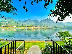 A view of a lake with mountains in the background, a pathway leading to the water, and a fence on the left. - Carambola Bungalow (Phong Nha)