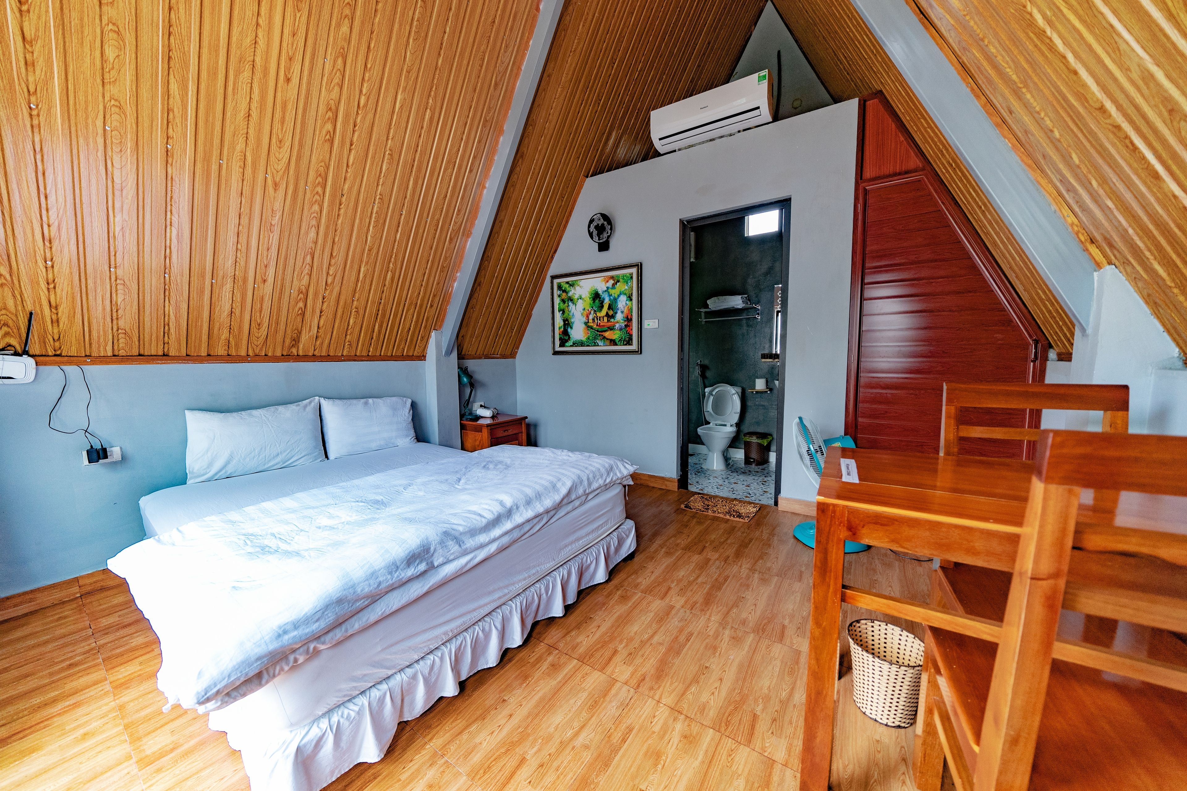 Un dormitorio con techo inclinado de madera, una cama, una silla de madera, una mesita y un aire acondicionado instalado en la pared.