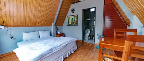 Un dormitorio con techo inclinado de madera, una cama, una silla de madera, una mesita y un aire acondicionado instalado en la pared.