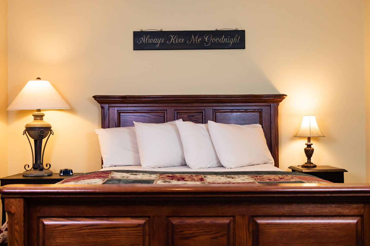Ein ordentlich bezogenes Bett mit Kopfteil, zwei Nachttischlampen und einem Schild über dem Bett mit der Aufschrift 'Always Kiss Me Goodnight'.