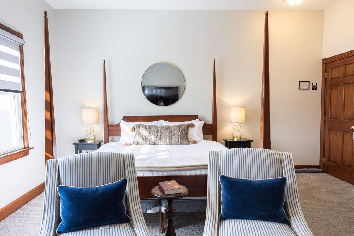 Ein Hotelzimmer mit einem großen Bett, zwei gestreiften Sesseln, einem Deckenventilator und einem runden Spiegel an der Wand.