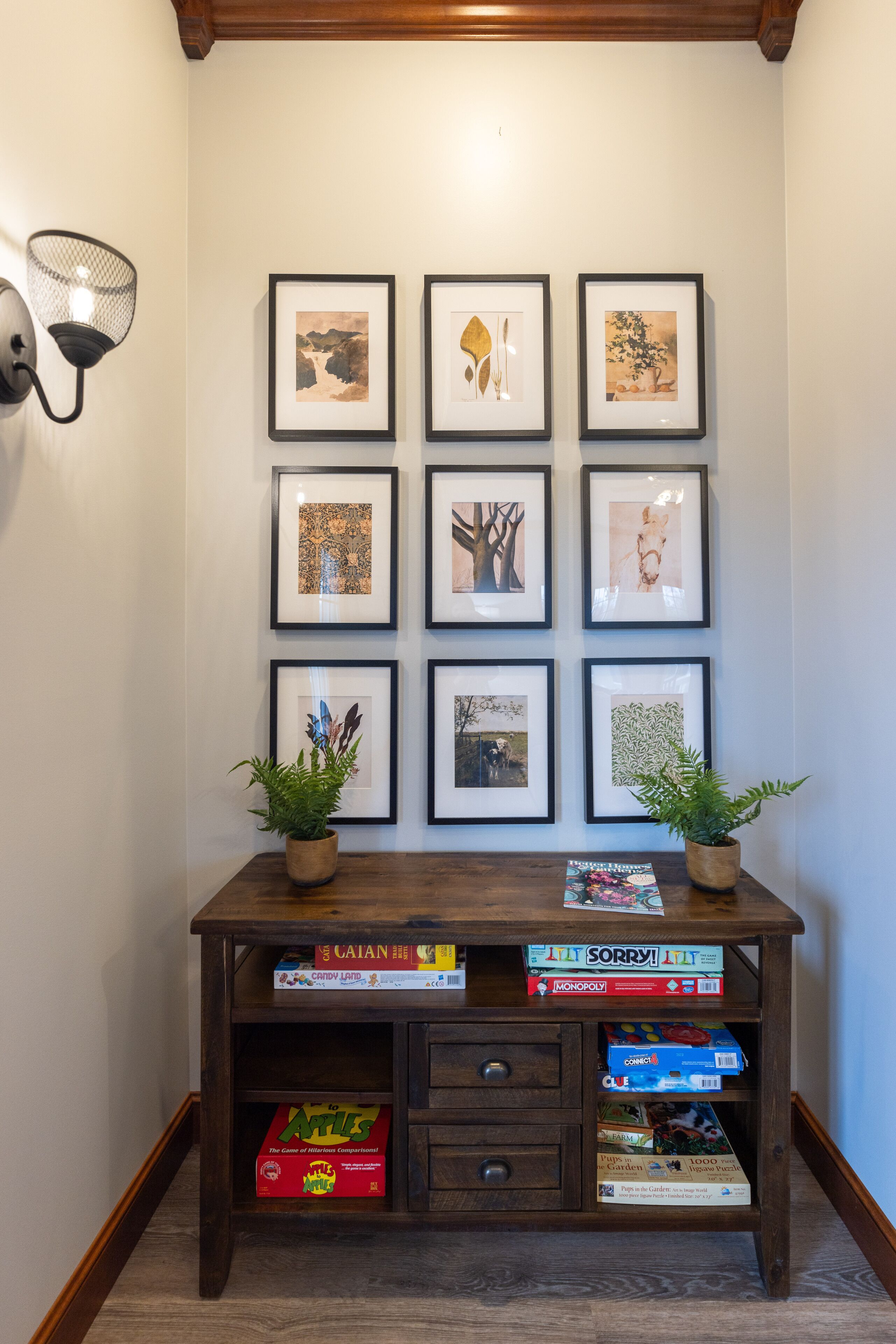 A hallway with framed artwork on the wall and a wooden console table with board games and potted plants.