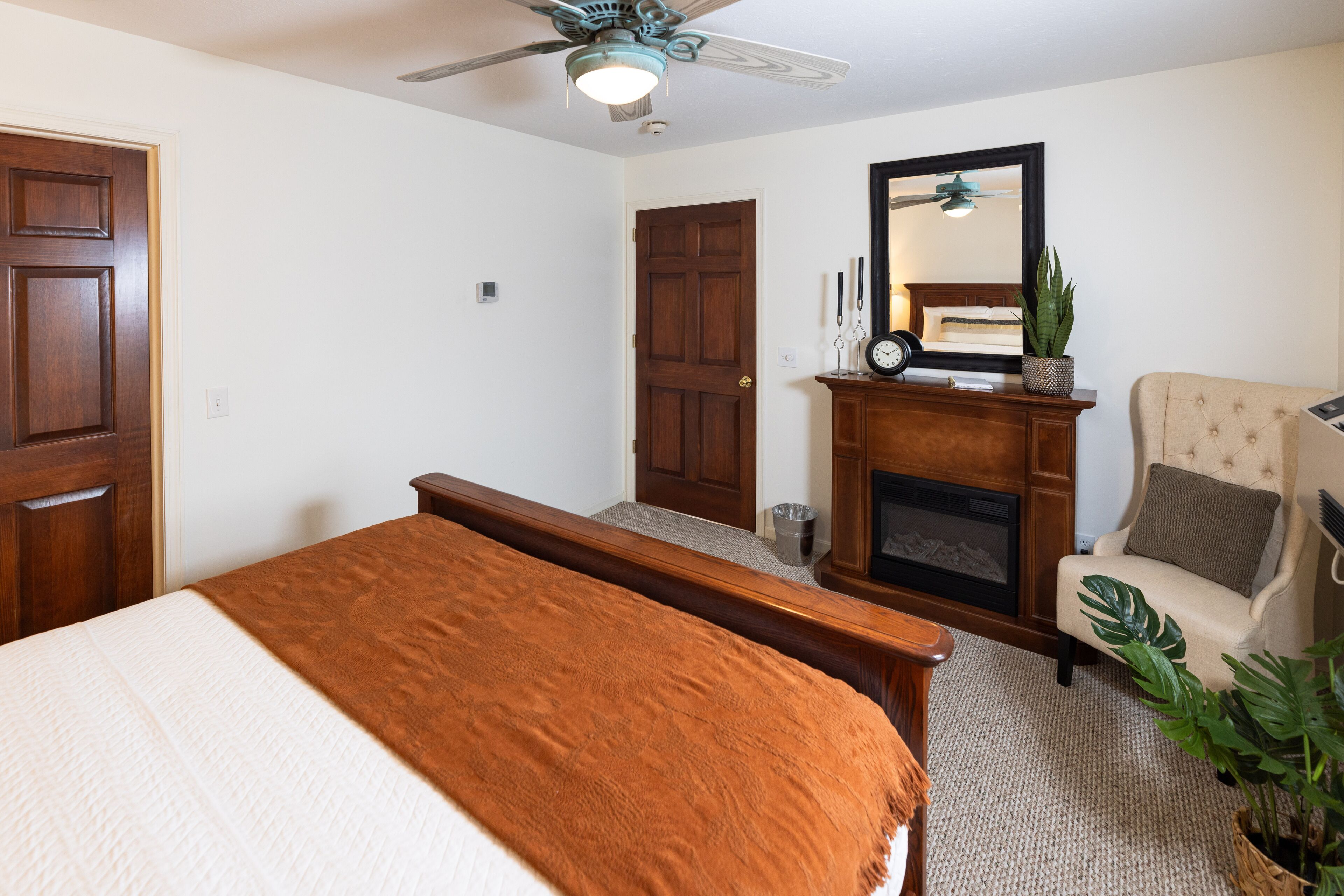 A bedroom with a bed, a wooden dresser, a chair, a fireplace, and a ceiling fan.