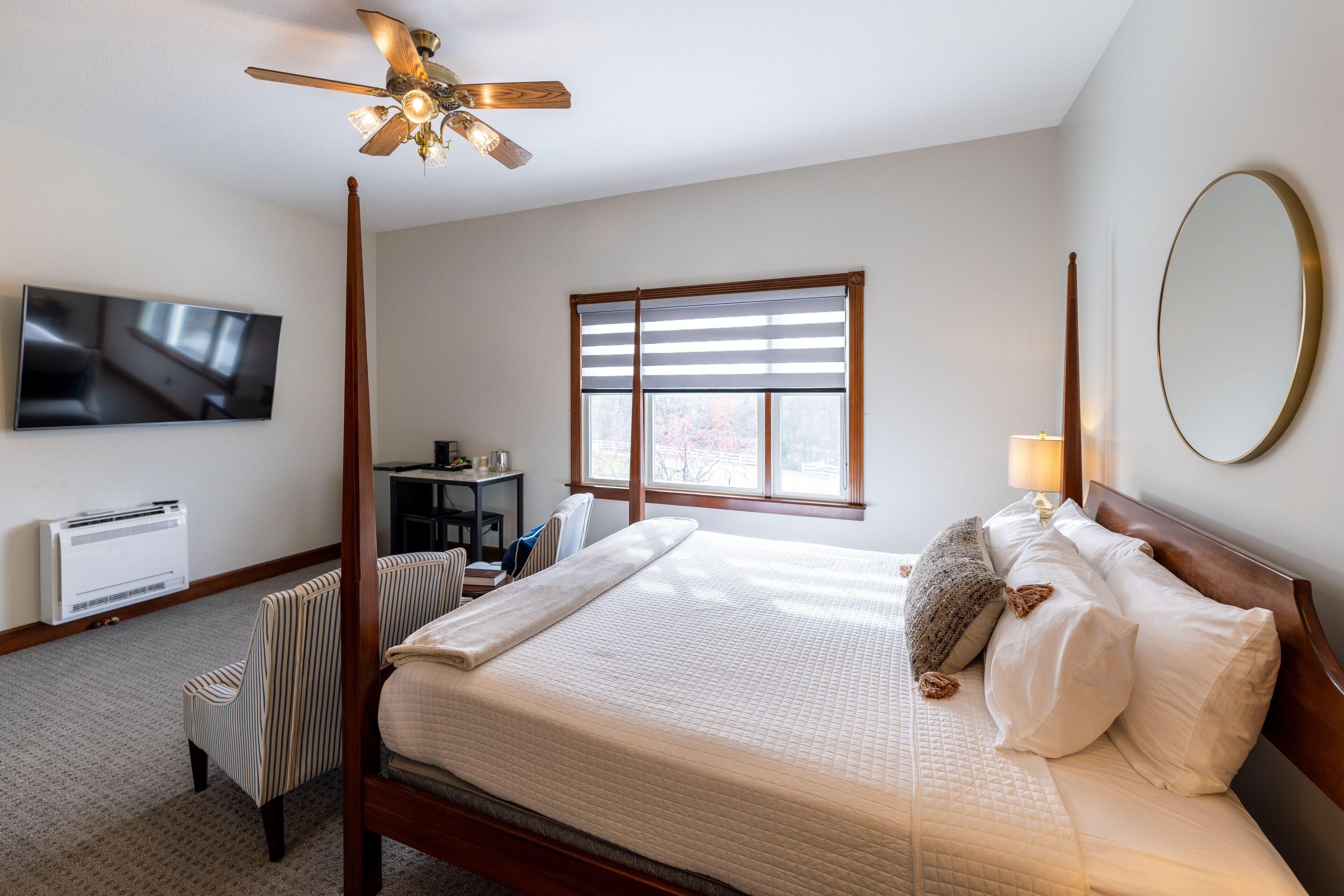A bedroom with a bed, a ceiling fan, a TV, a window with blinds, a chair, and a bedside table.