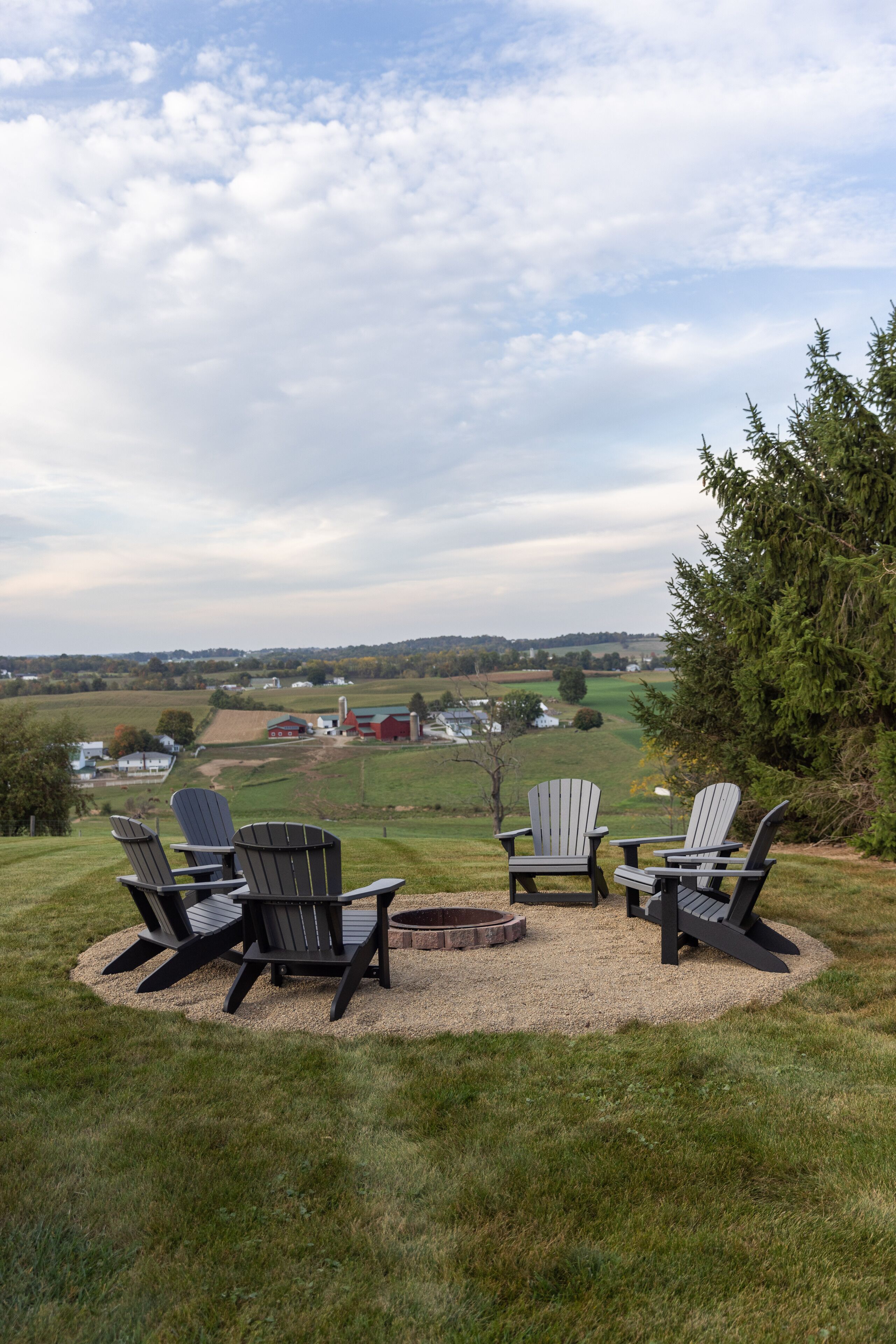 A circular seating area with Adirondack chairs and a fire pit, overlooking a rural landscape with fields and a distant red barn.