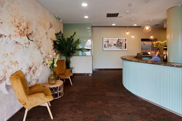 A reception area with a curved counter, a large wall with a floral mural, and a seating area with two chairs and a small table. - Ramblas Vendrell (El Vendrell)