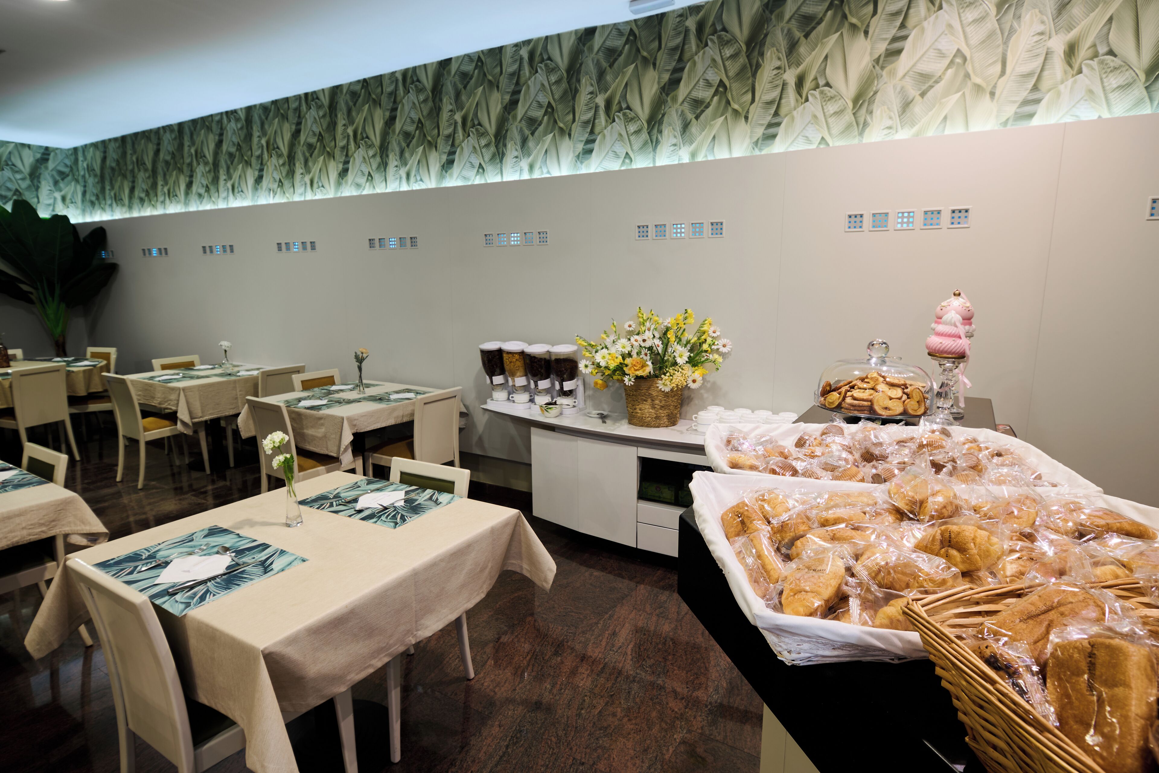 Un comedor con mesas dispuestas para el servicio, un bufé con diversas opciones de comida y una pared decorativa con motivos de hojas.