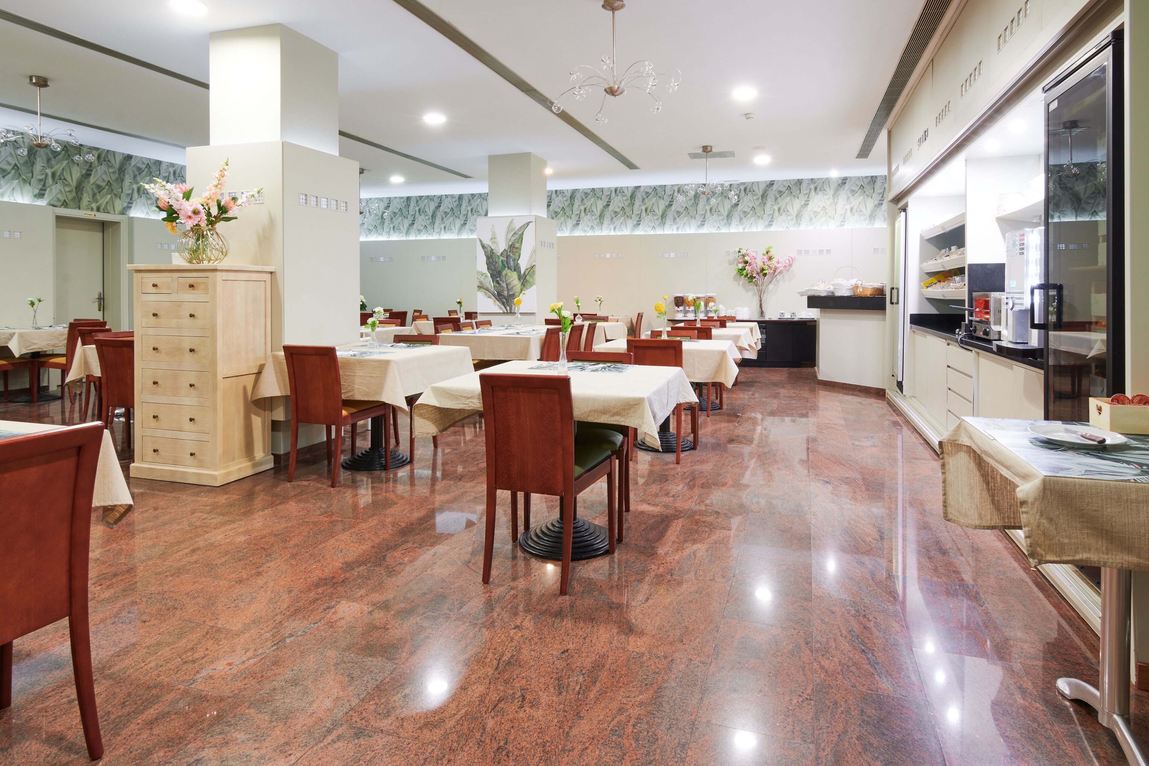 A spacious restaurant with multiple tables and chairs, a kitchen area, and a decorative plant arrangement.