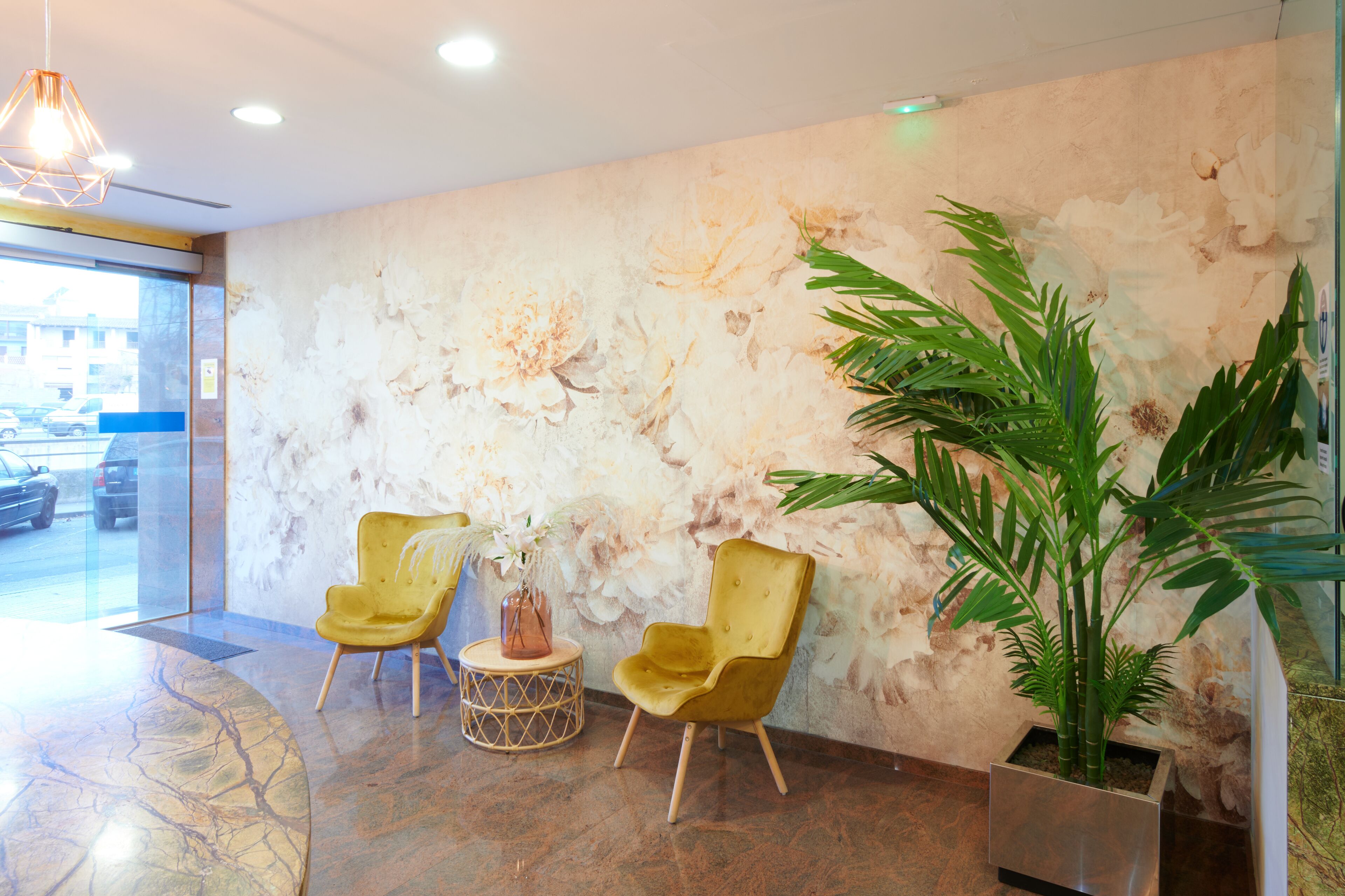 Un área de recepción con dos sillas amarillas, una planta grande en maceta y una pared con un mural floral.