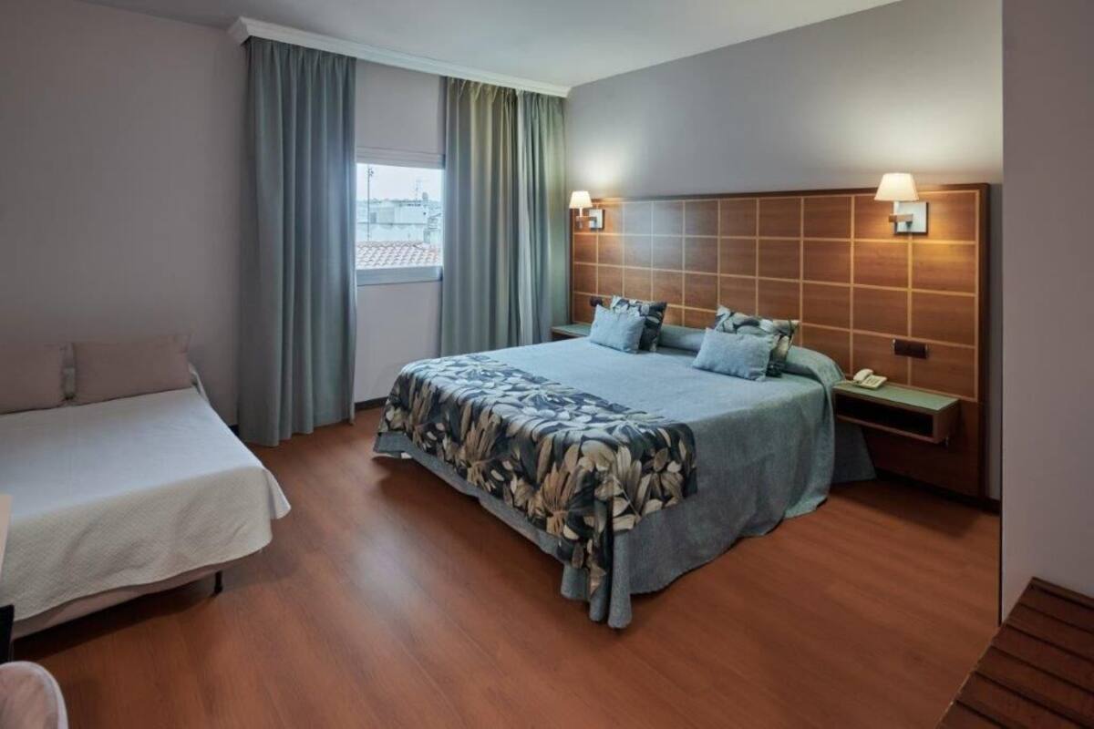 Habitación de hotel con una cama grande, una cama más pequeña, una mesita de noche con lámpara y una ventana con cortinas.
