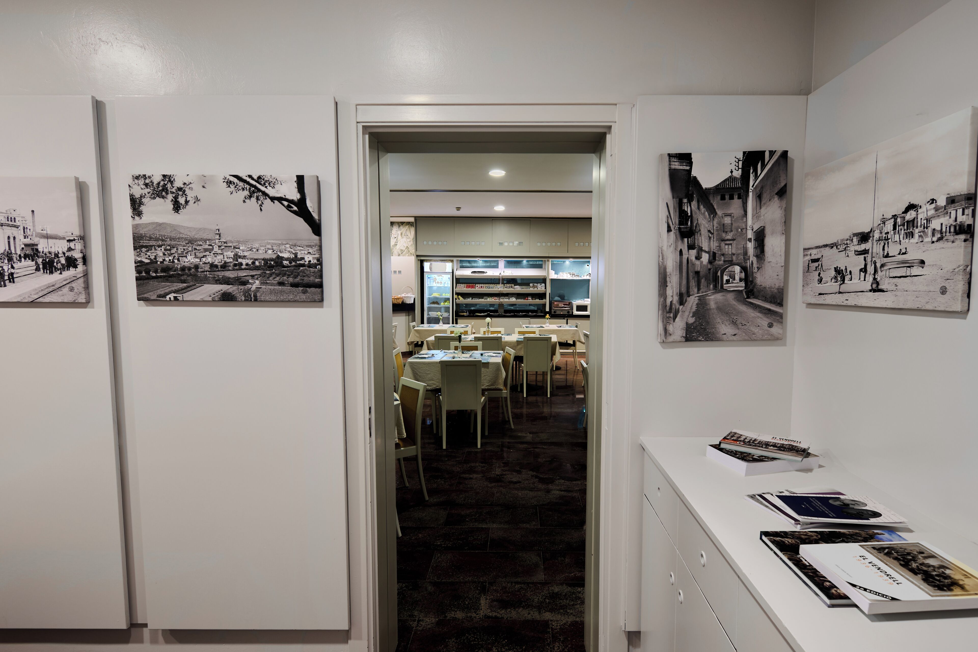 A hallway with framed black and white photographs on the walls, leading to a dining area with tables and chairs.