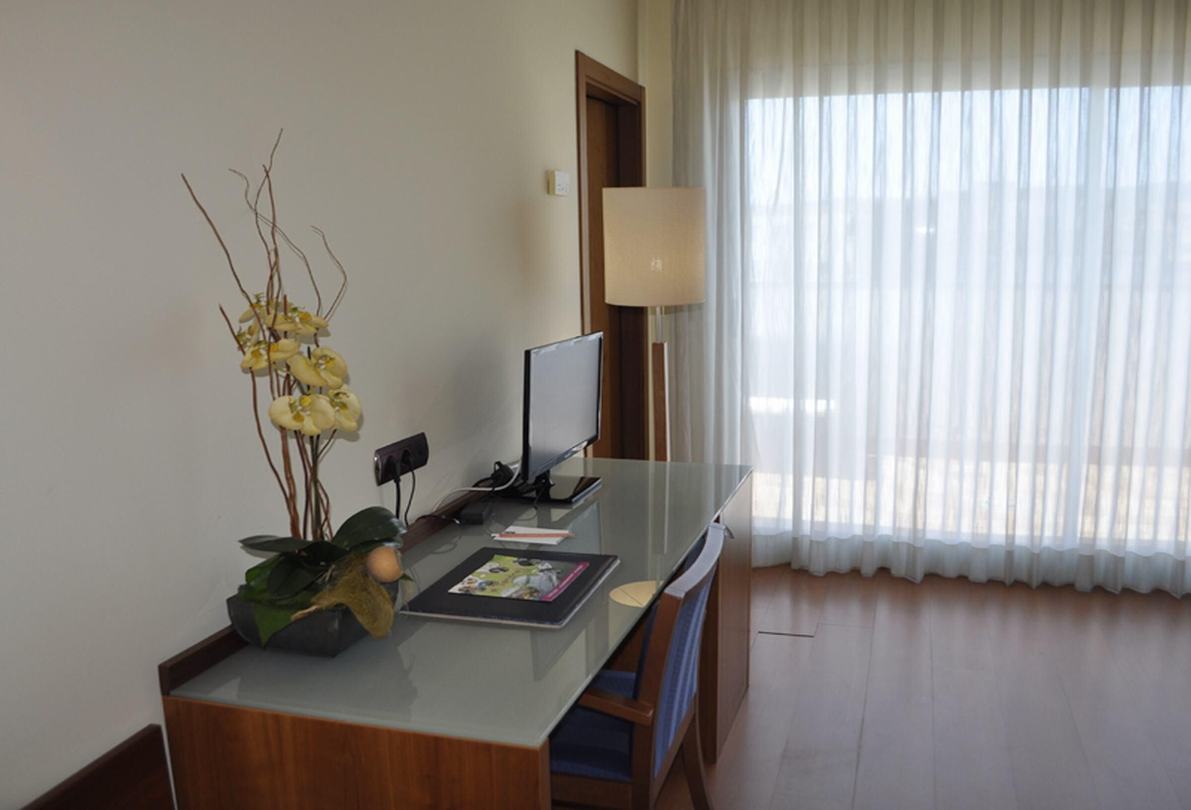 A hotel room with a glass desk, a television, a lamp, and a window with sheer curtains.