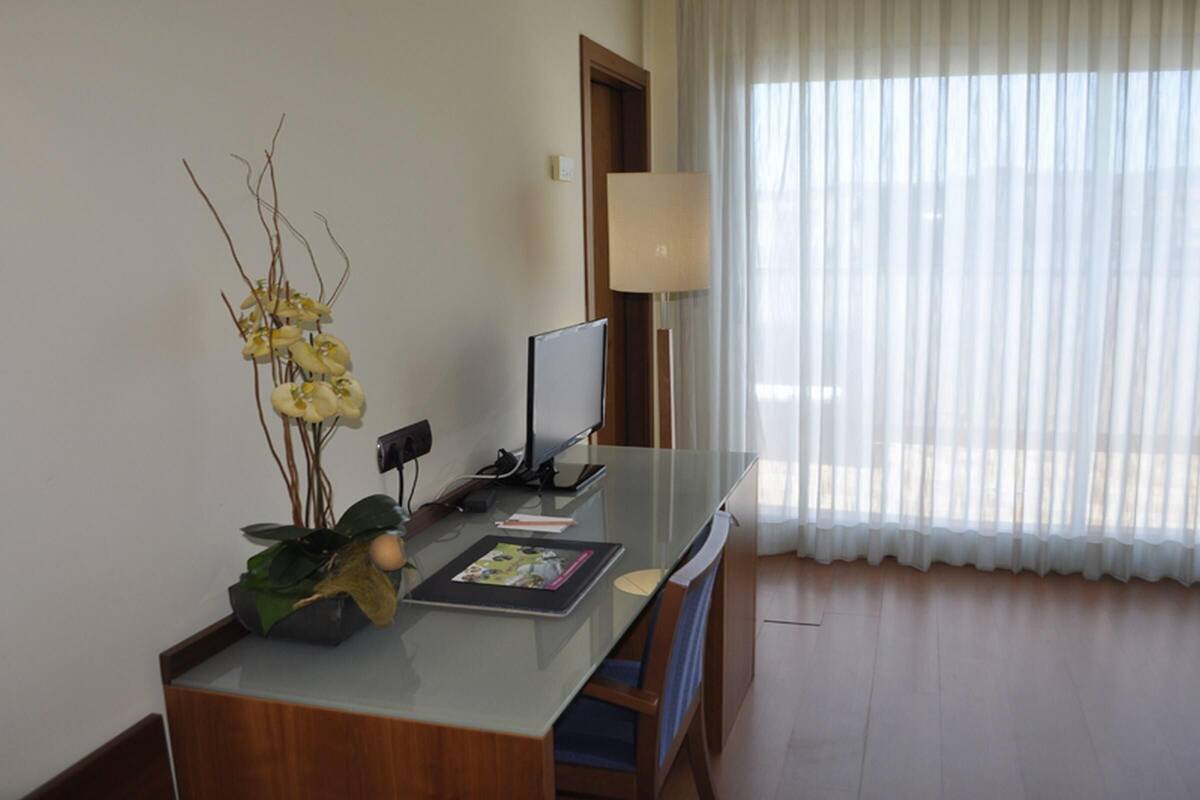 Ein Hotelzimmer mit Glastisch, Fernseher, Lampe und einem Fenster mit transparenten Vorhängen.