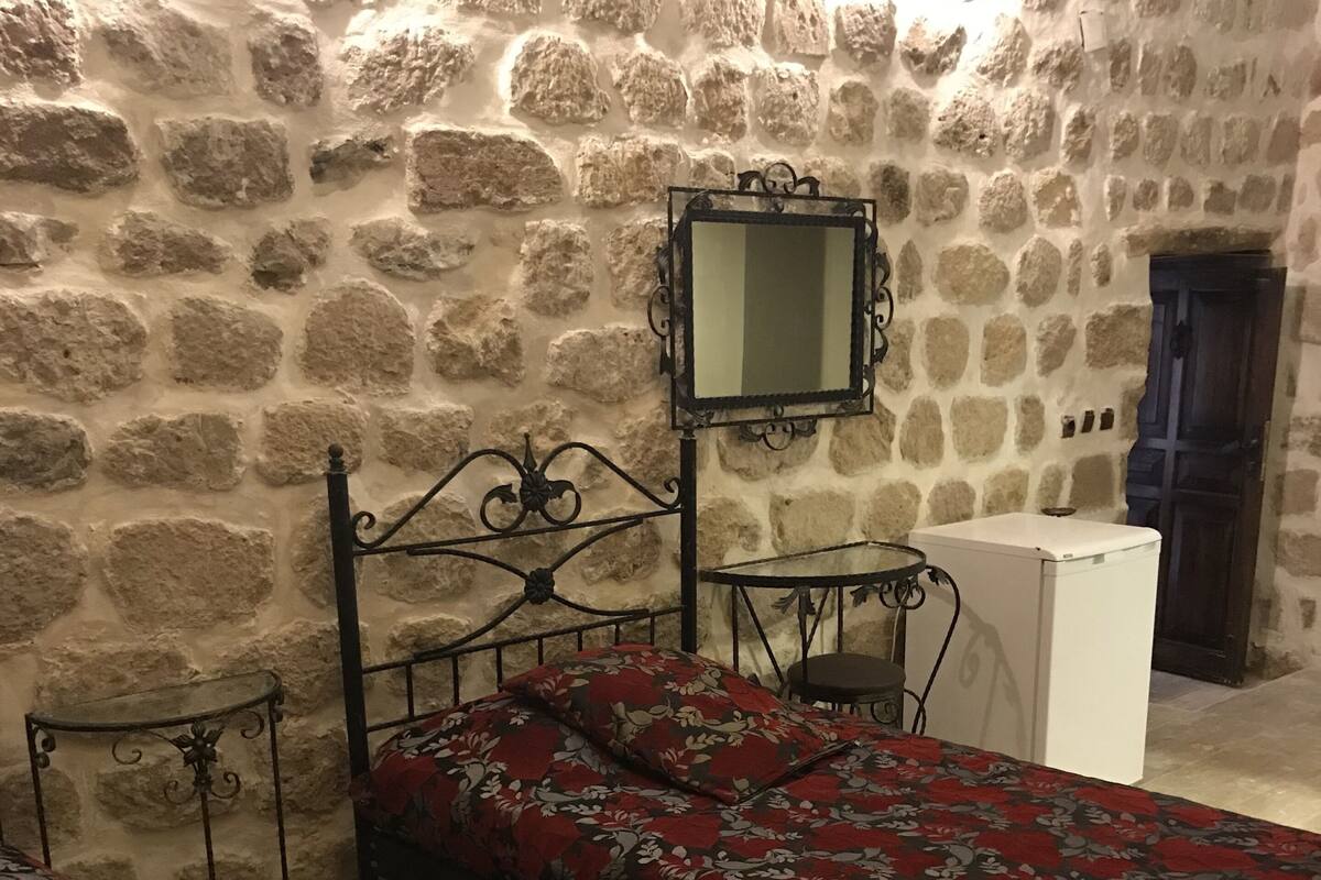 A bedroom with a stone wall, a bed with a floral bedspread, a small table, and a mirror.