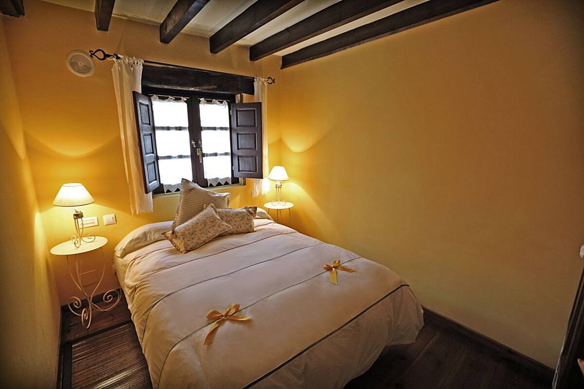 A bedroom with a bed, two bedside lamps, a window with curtains, and a wooden ceiling.