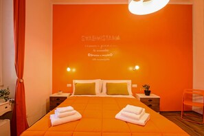 A hotel room with an orange theme, featuring a bed with two pillows, a nightstand with a plant, and a chair. - Allegras House Caracciolo (Rome)
