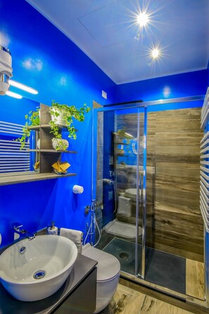 A modern bathroom with a blue wall, a white sink, a glass shower enclosure, and a wooden floor. - Allegras House Caracciolo (Rome)