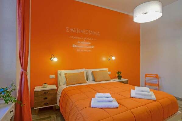 A bedroom with an orange wall, a bed with a white and orange bedspread, two bedside tables with lamps, and a small potted plant. - Allegras House Caracciolo (Rome)