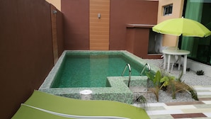 Una piscina petita a l'aire lliure amb escales de rajola, una cadira relaxant verda, una taula blanca amb un para-sol groc i una petita zona de jardí amb plantes i pedres.