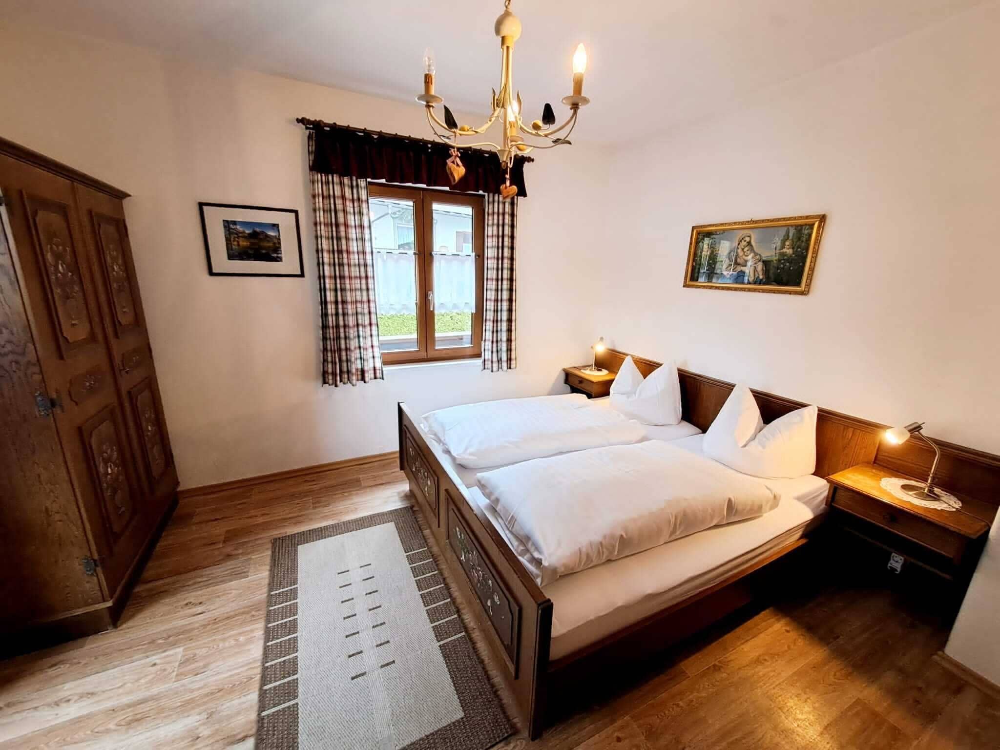 A bedroom with a wooden bed, a chandelier, a window with curtains, a painting, and a wardrobe.
