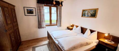 A bedroom with a wooden bed, a chandelier, a window with curtains, a painting, and a wardrobe.