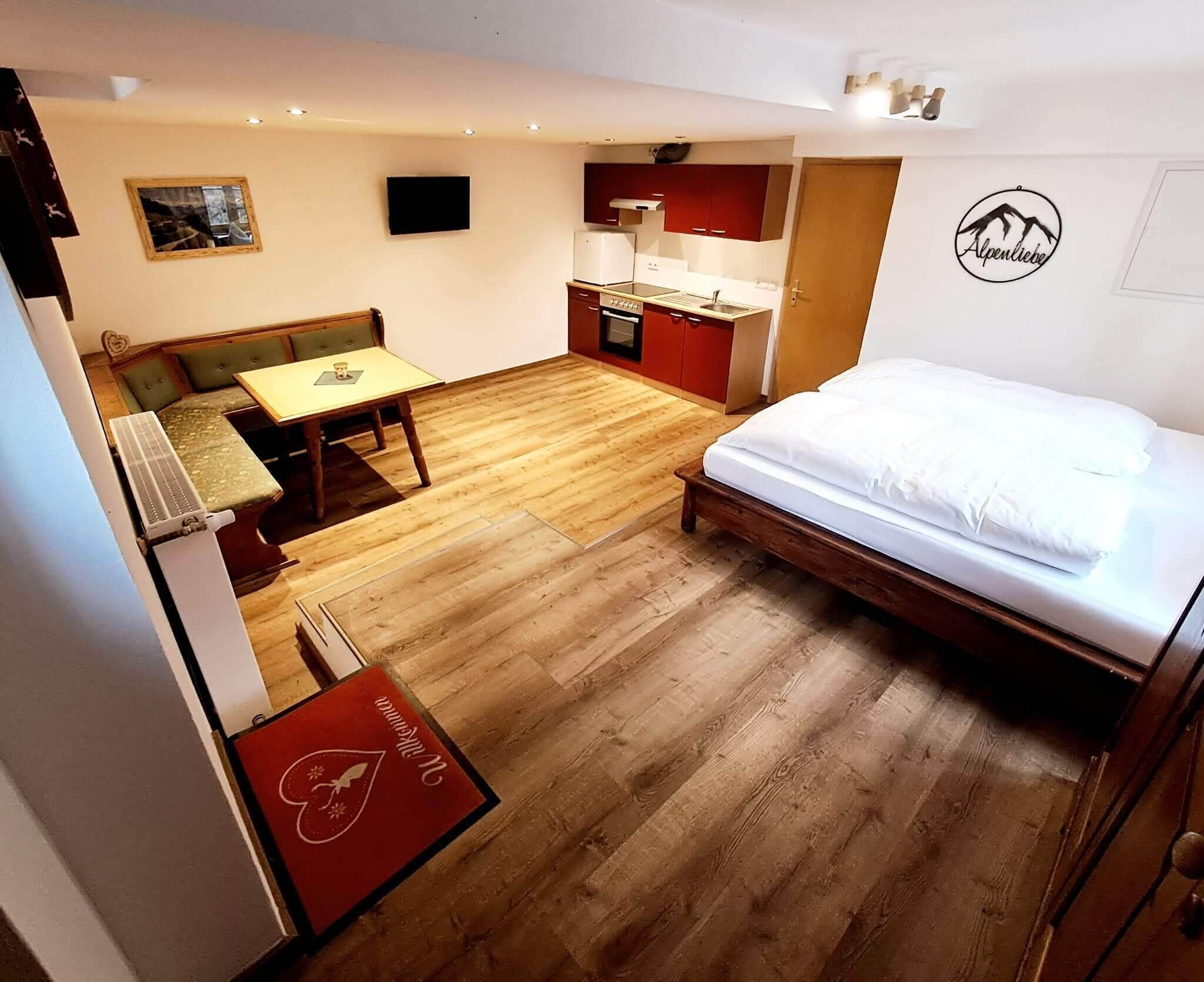 A compact hotel room with a wooden floor, a bed, a small kitchenette, a dining area with a table and chairs, and a TV mounted on the wall.