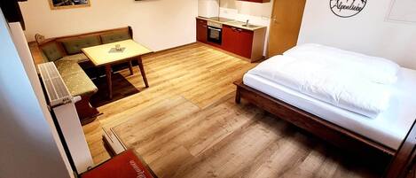 A compact hotel room with a wooden floor, a bed, a small kitchenette, a dining area with a table and chairs, and a TV mounted on the wall.