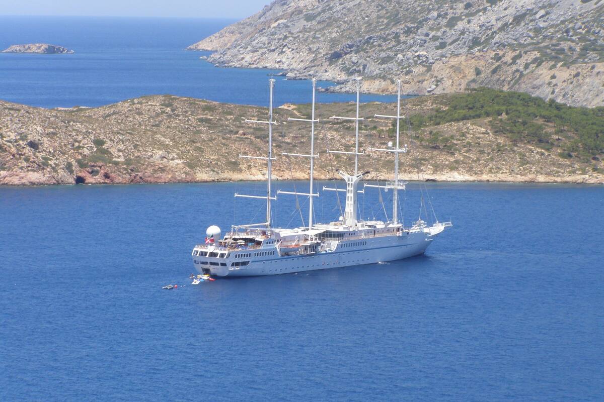 A large sailing ship on calm waters with rocky terrain and a clear sky in the background.