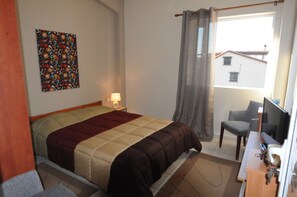 Ett sovrum med en säng, ett skrivbord med en dator, en stol, ett fönster med utsikt över byggnader och en vägg med ett färgstarkt konstverk.