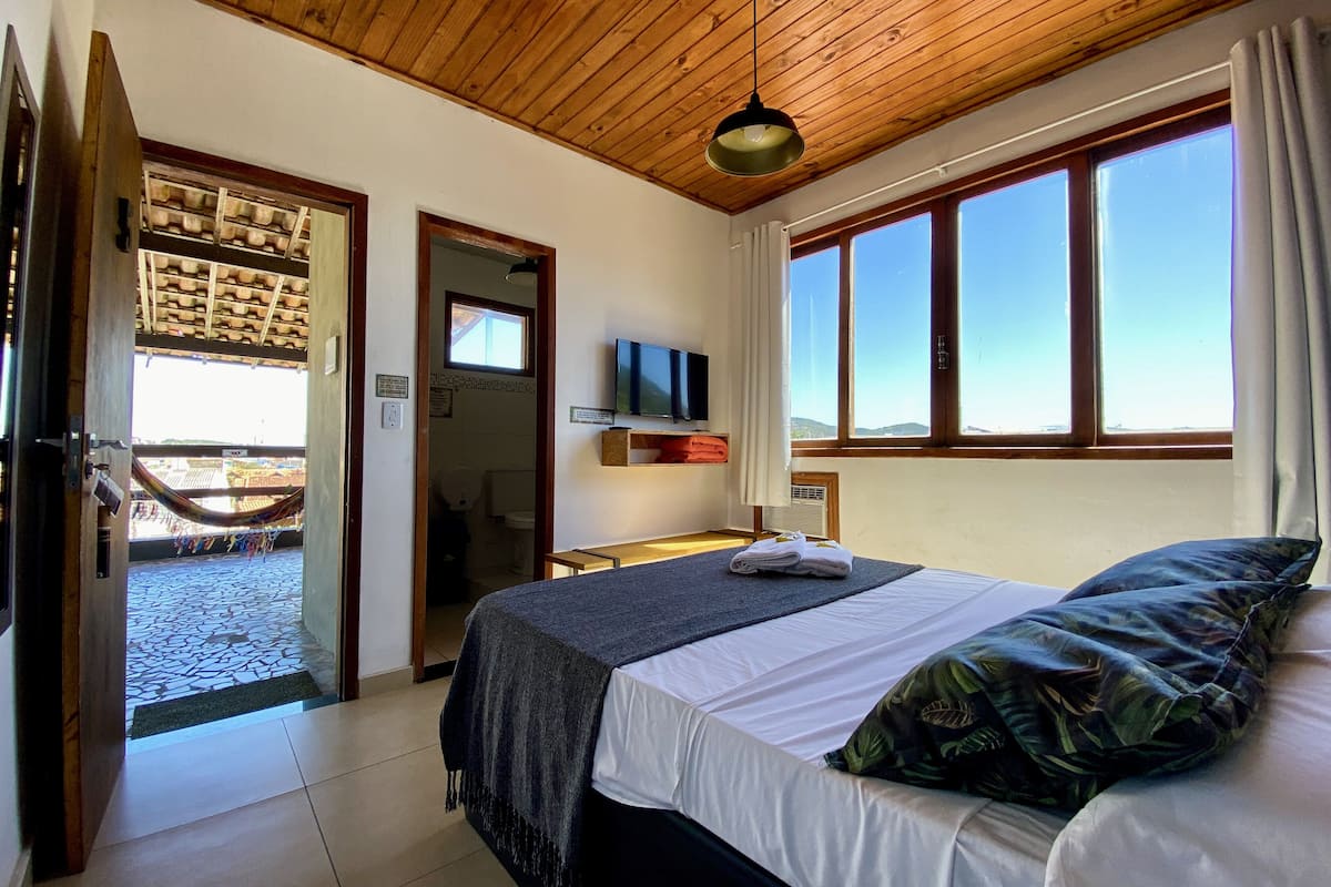 A hotel room with a bed, a TV, a window with curtains, a wooden ceiling, and a view of the outside.