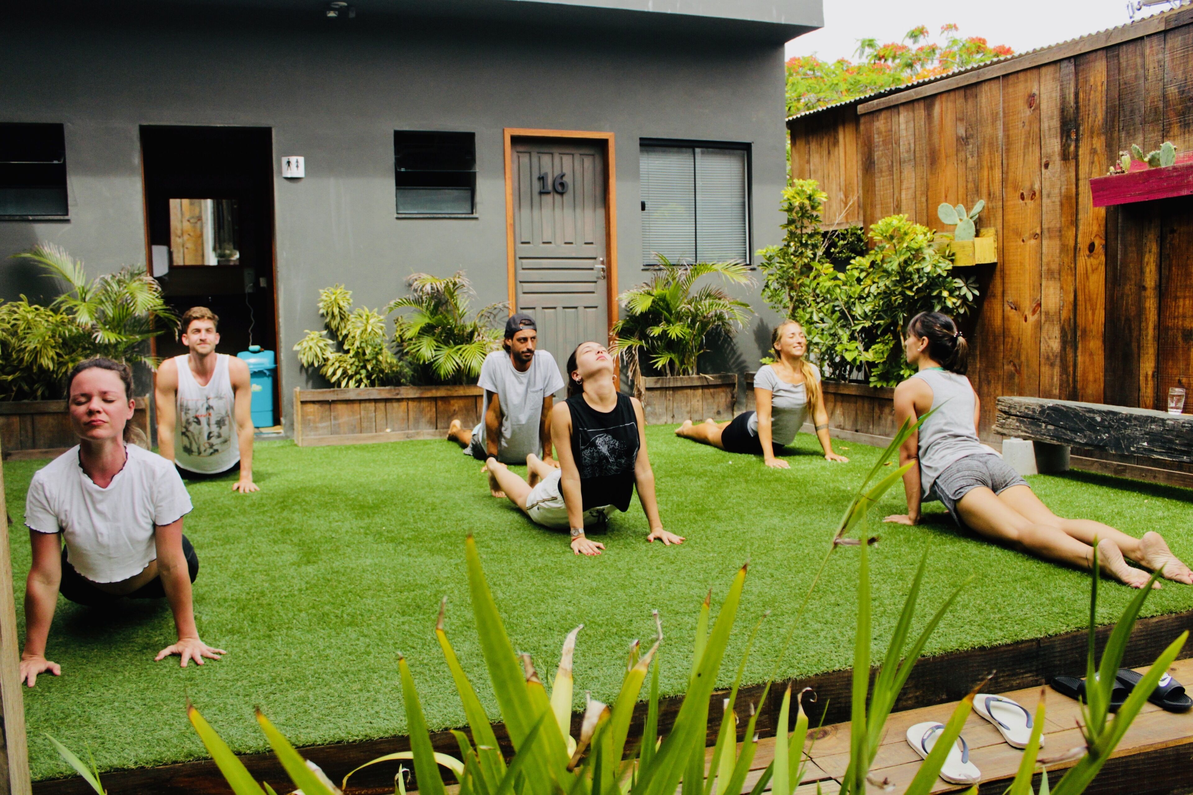 A group of people doing a workout on artificial grass in an outdoor setting with wooden fencing and potted plants.