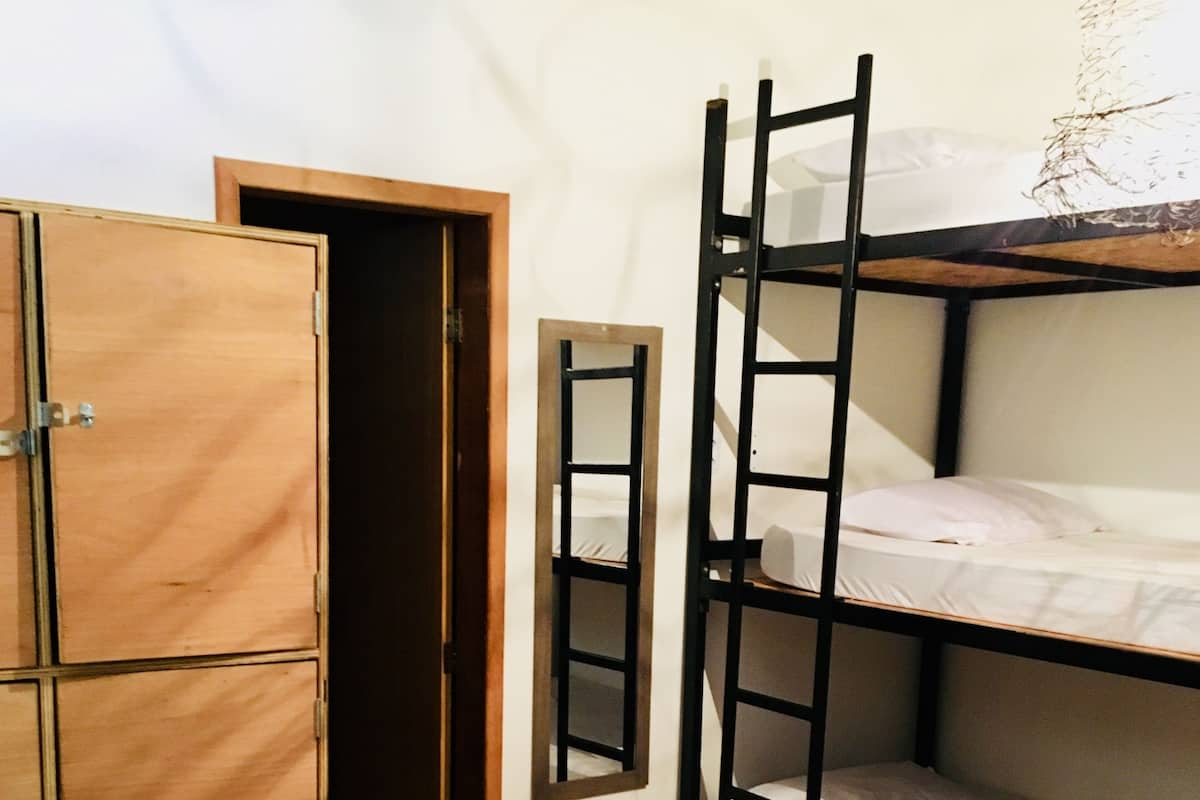 A dormitory room with bunk beds, a wooden wardrobe, and a door leading to another room.