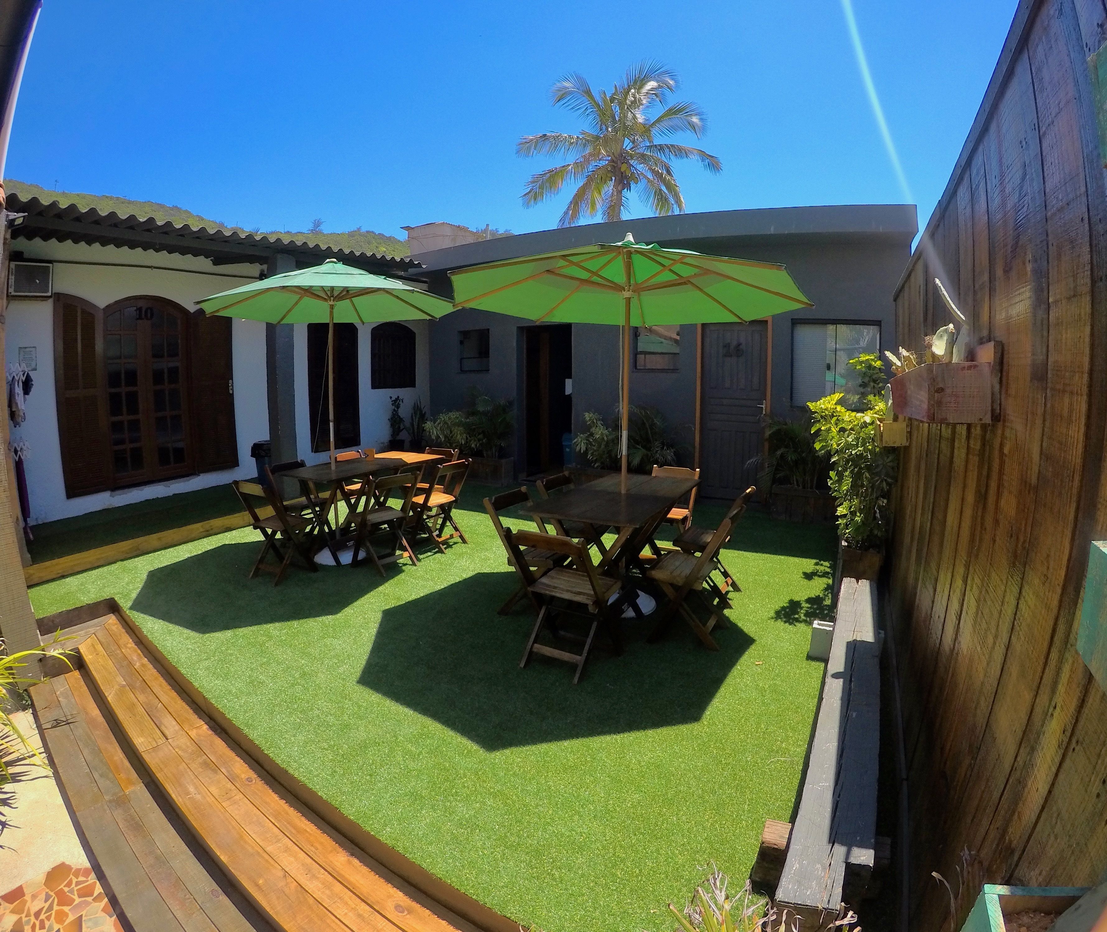 An outdoor patio with wooden tables and green umbrellas, surrounded by a wooden fence and a palm tree.