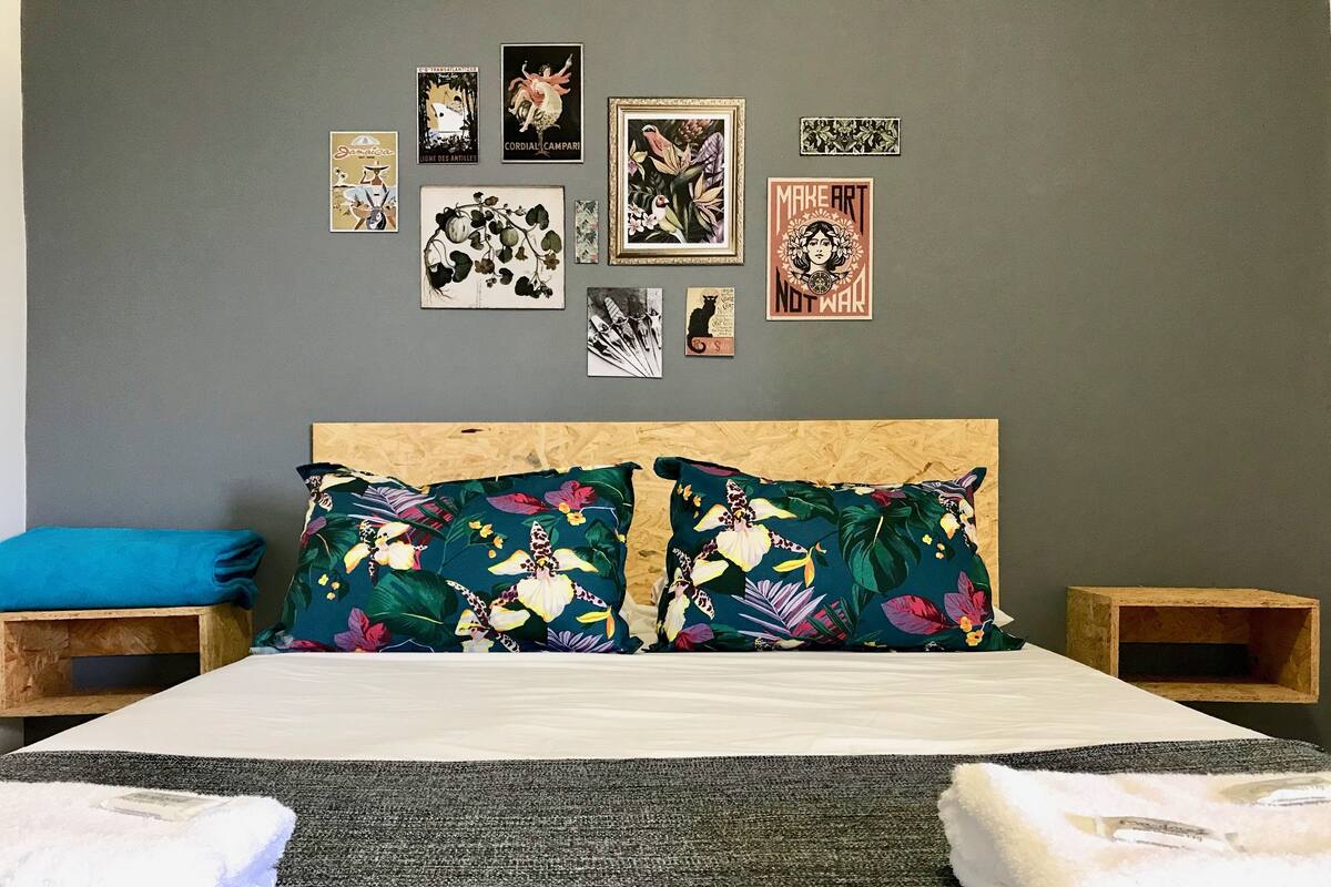 A bedroom with a bed, floral pillows, a grey blanket, and artwork on the wall.