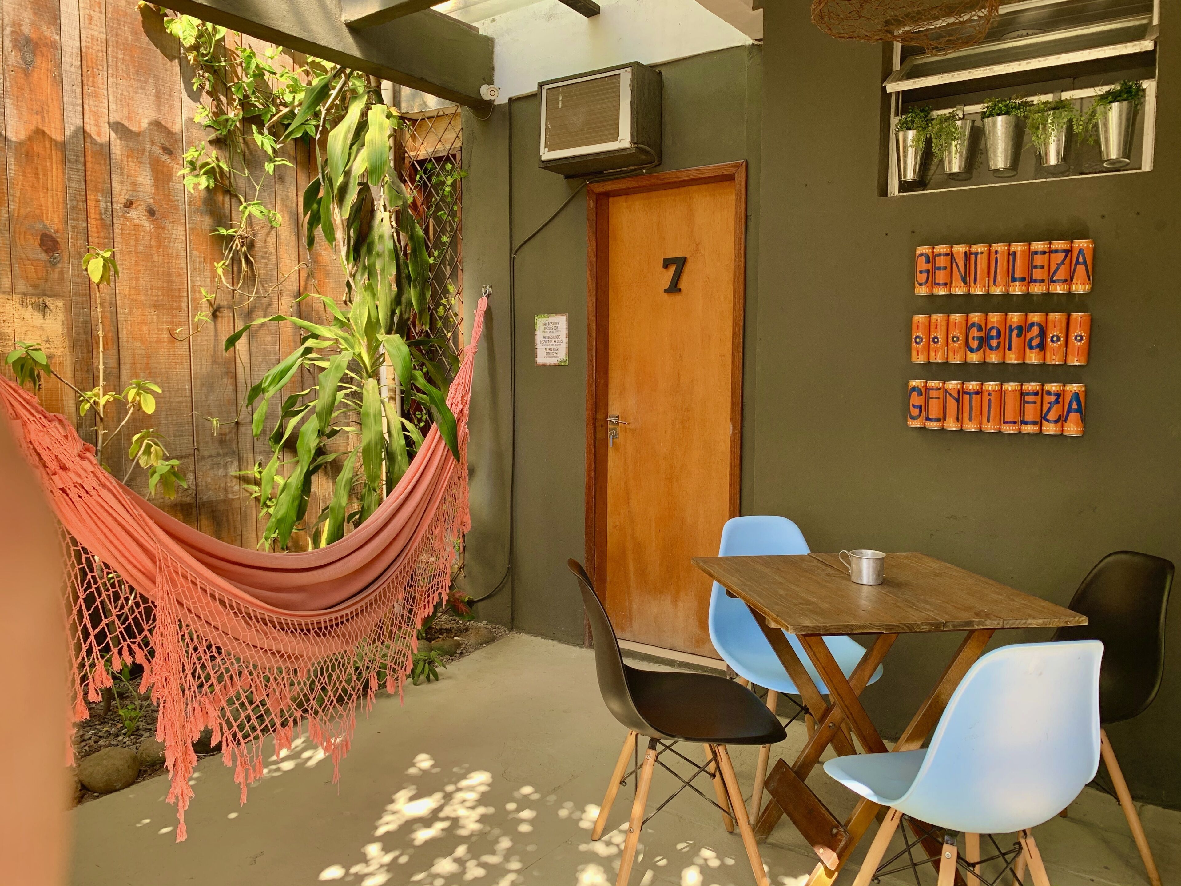 Una habitación con puerta de madera marcada con el número 7, una hamaca, una mesa con sillas y una pared con plantas en macetas y un cartel que dice 'GENTILEZA'.