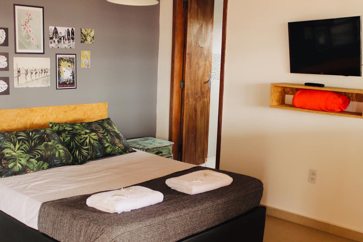 A neatly made bed with a grey headboard and tropical leaf-patterned pillows, a flat-screen TV mounted on the wall, and a wooden shelf with a red pillow.