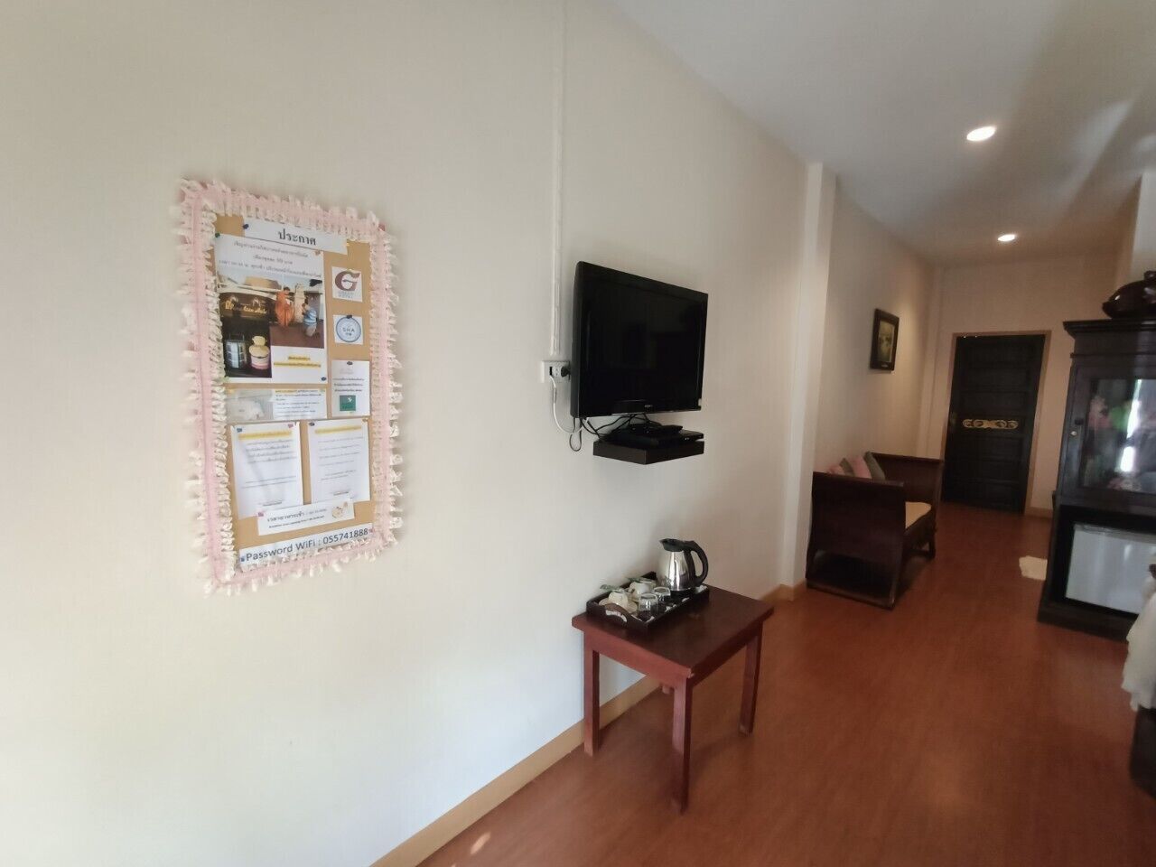 A hallway with a bulletin board, a television mounted on the wall, a small table with a kettle, and a wooden chair.