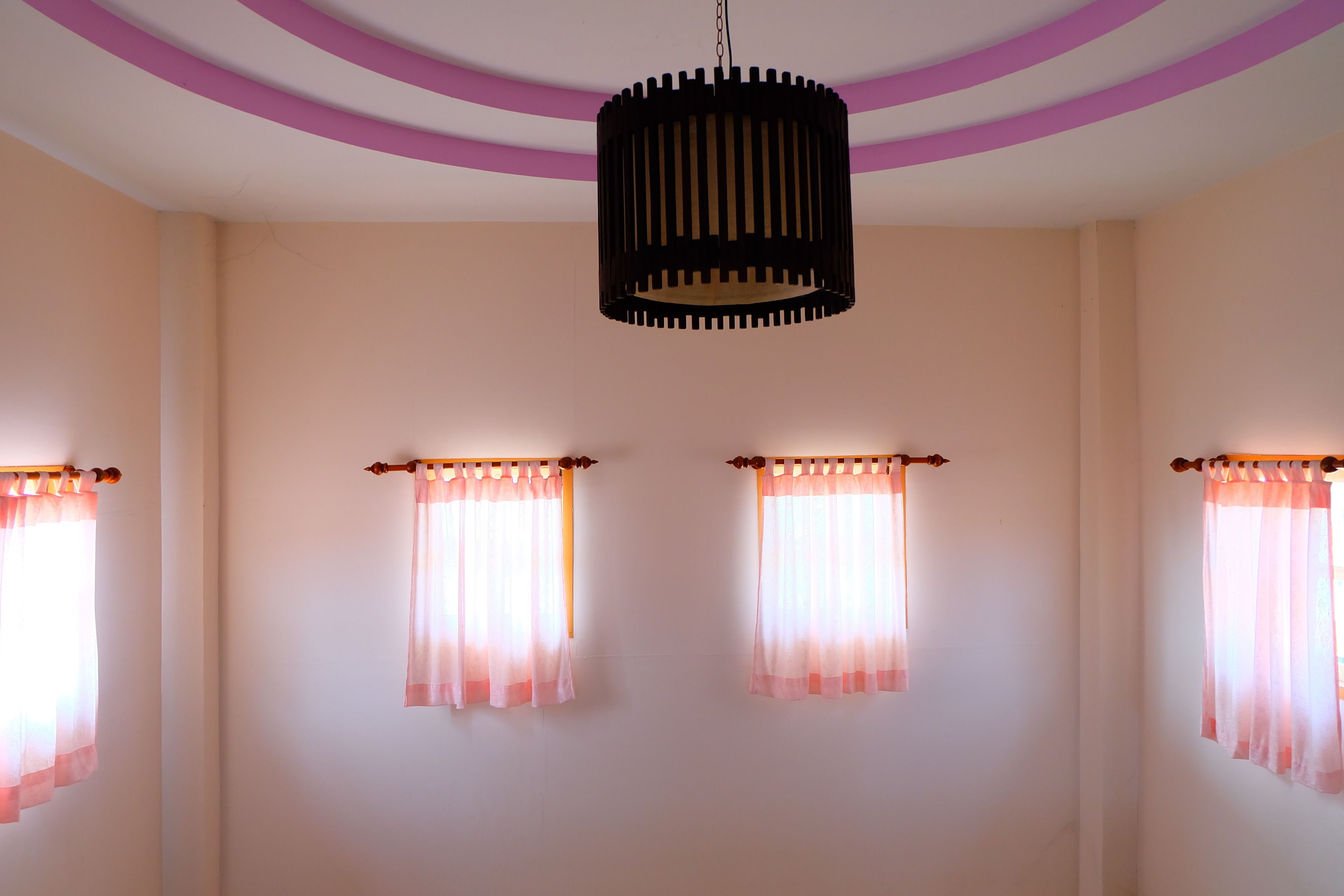 A room with a ceiling lamp, two windows with curtains, and a pink and white wall design.