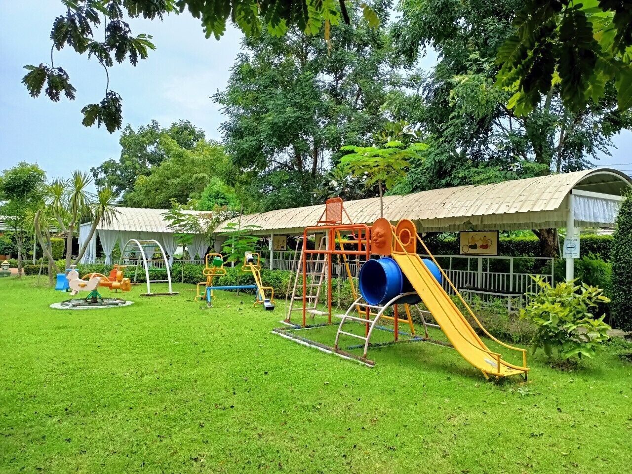 A playground with various play structures including slides and swings, surrounded by lush greenery and trees.
