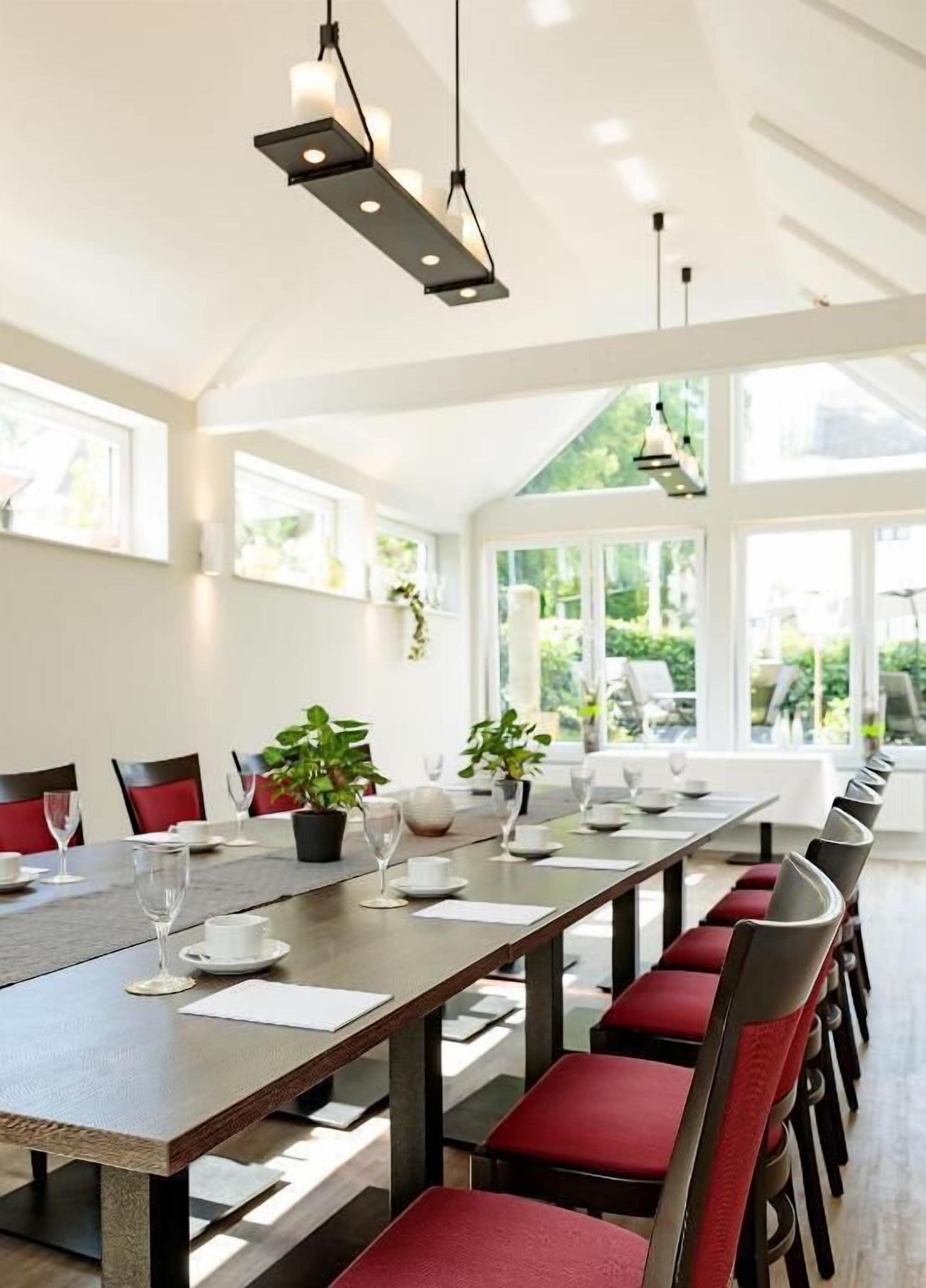 A long dining table with chairs, set for a meal, in a bright room with large windows and natural light.