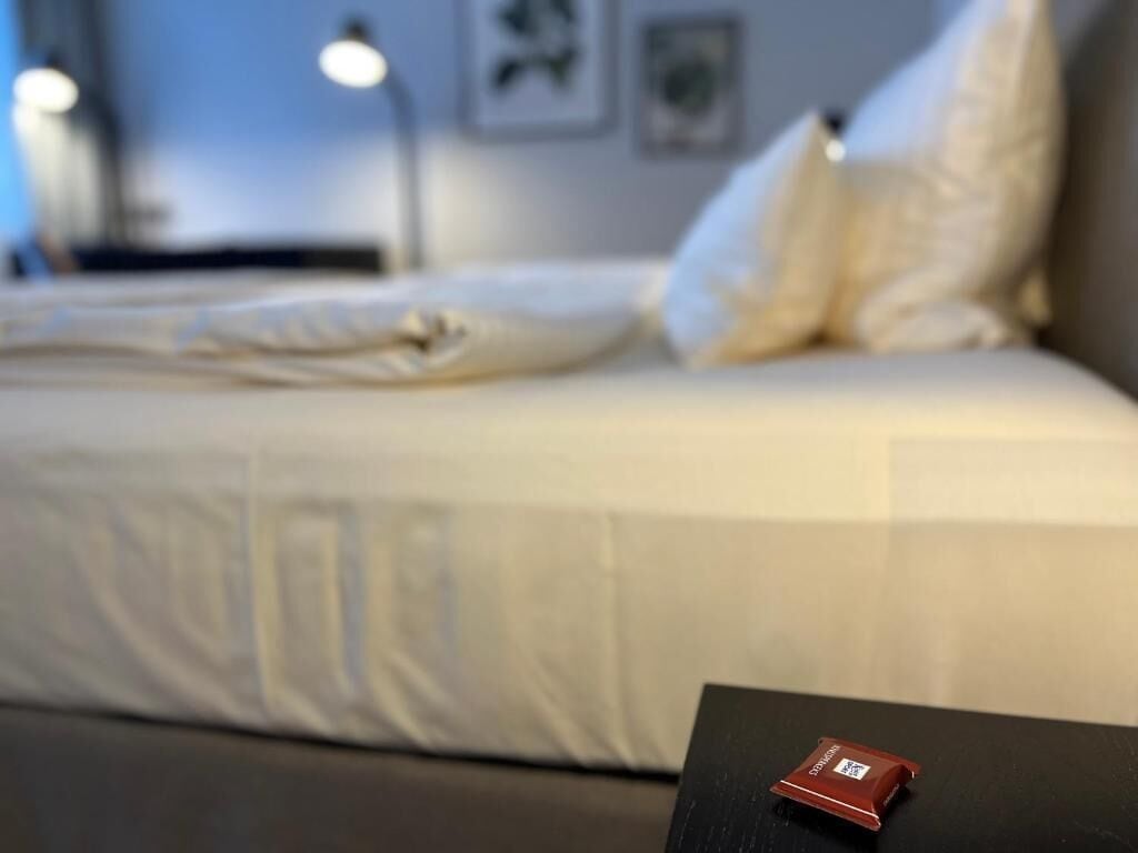 Ein Bett mit weißer Bettwäsche und Kissen, ein schwarzer Tisch mit einem roten Gegenstand darauf und eine Wand mit gerahmten Bildern.