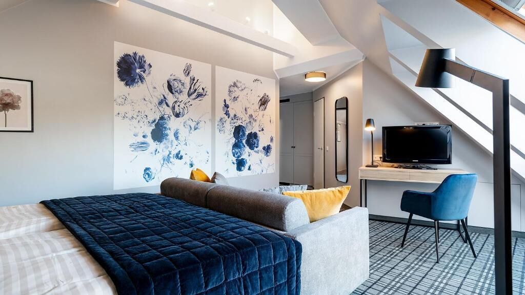 A hotel room with a bed, a desk with a TV, a chair, and artwork on the wall.