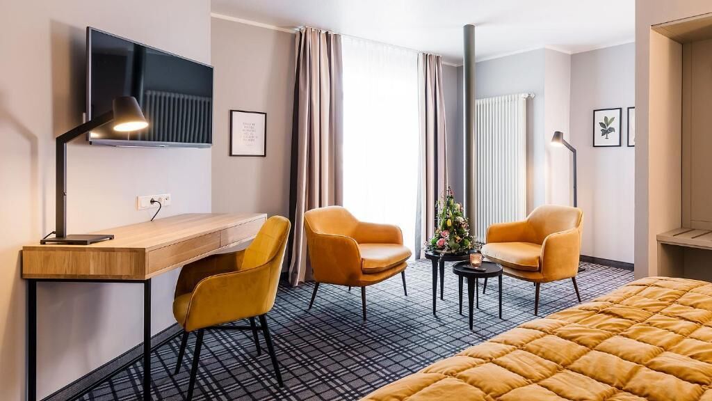 A hotel room with a bed, two chairs, a desk, a television, and a radiator.
